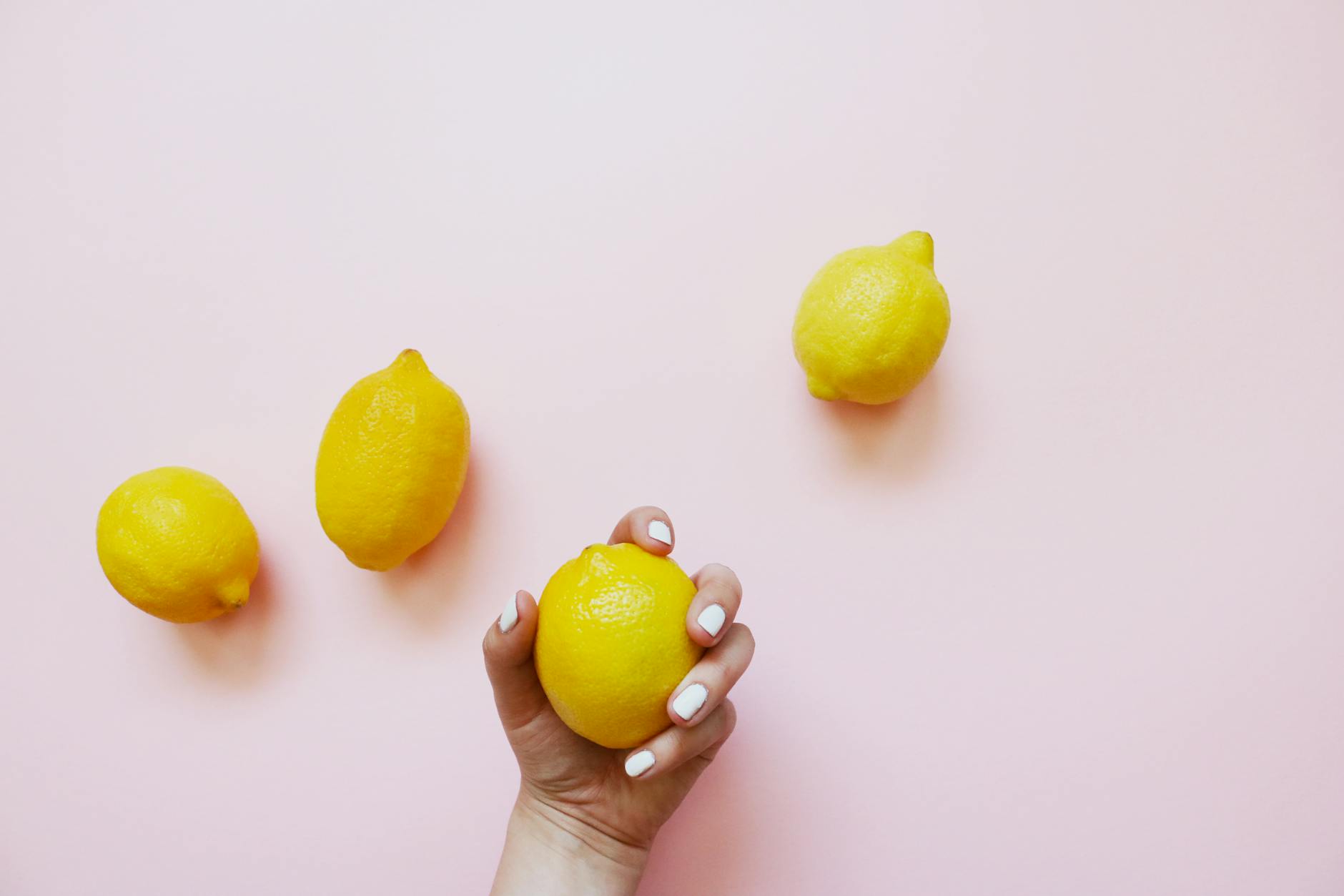 A hand holding a lemon against a soft pink background, surrounded by fresh lemons symbolizing natural sensation and touch.