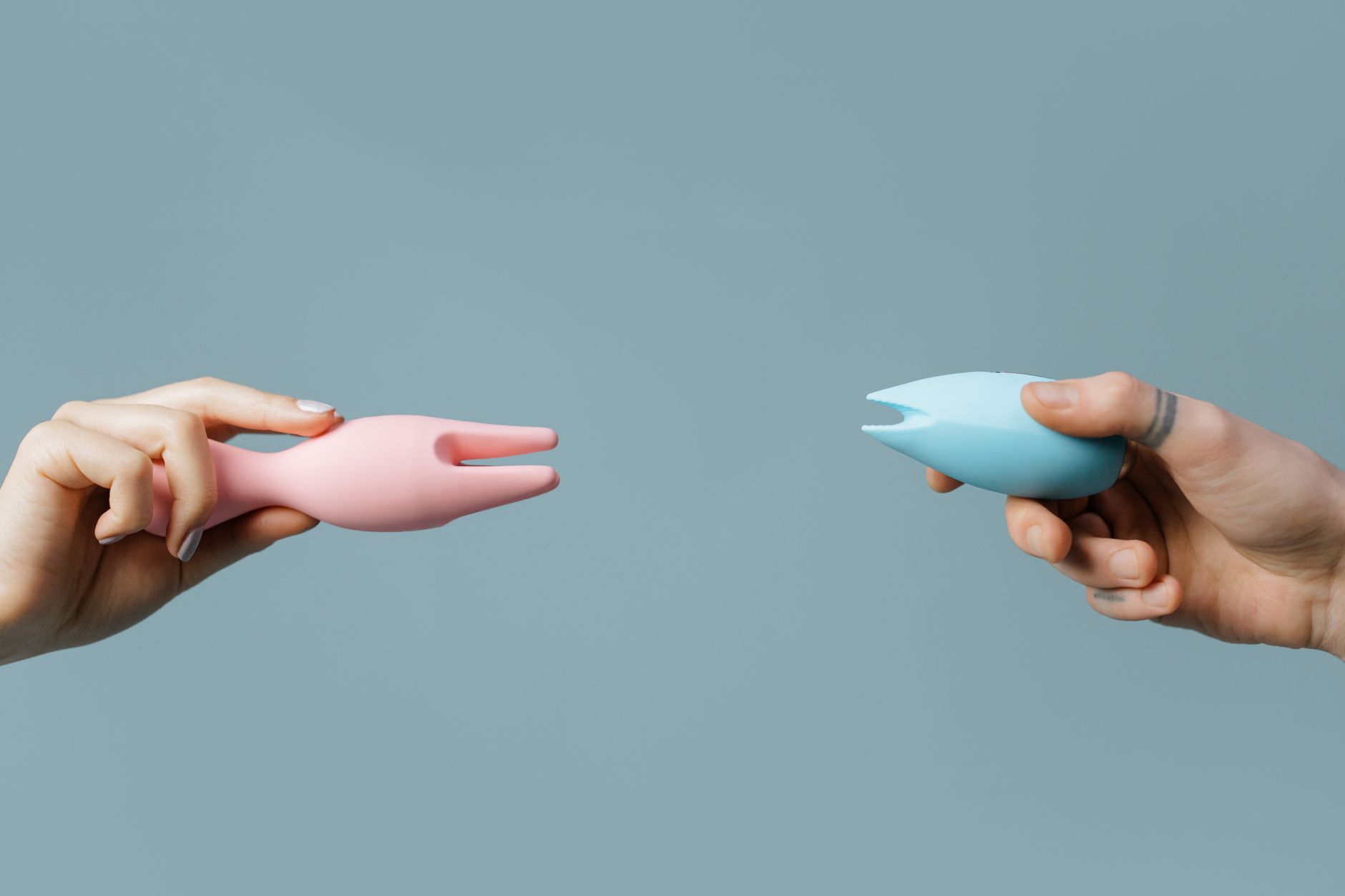 Two hands holding colorful vibrators against a soft pastel background, symbolizing shared pleasure and intimacy.
