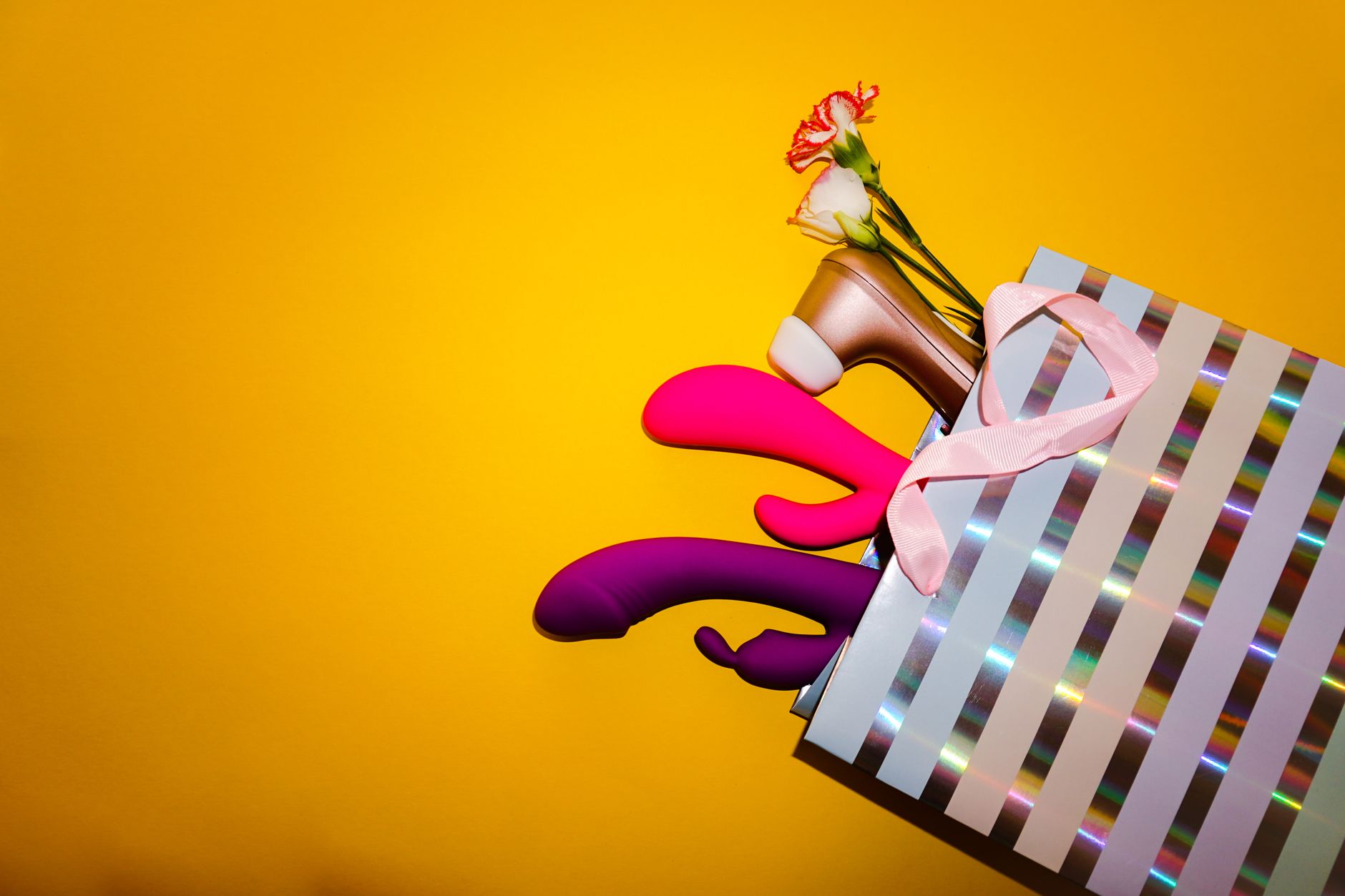 Colorful vibrators with fresh flowers in holographic packaging against a bright yellow background, representing pleasure variety and reset strategies